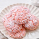 A stack of Strawberry Crinkle Cookies with white powdered sugar cracks on a white plate.